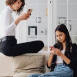 Two women using smartphones while seated on a couch in a modern living room setting.