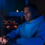 Man in hoodie types on laptop, illuminated by screen in dimly lit room with city lights.