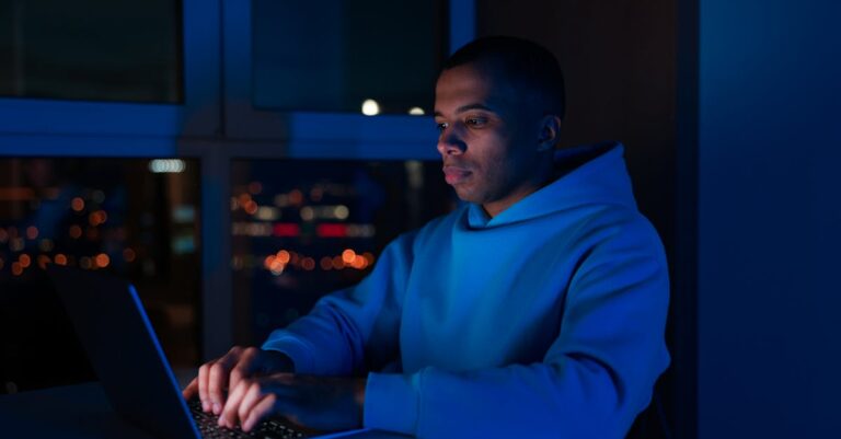 Man in hoodie types on laptop, illuminated by screen in dimly lit room with city lights.